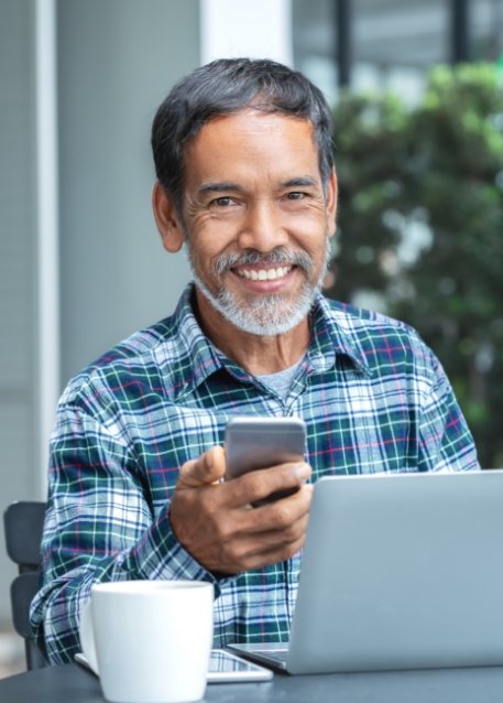 Man in blue plaid shirt holding his phone while sitting at a table with a laptop